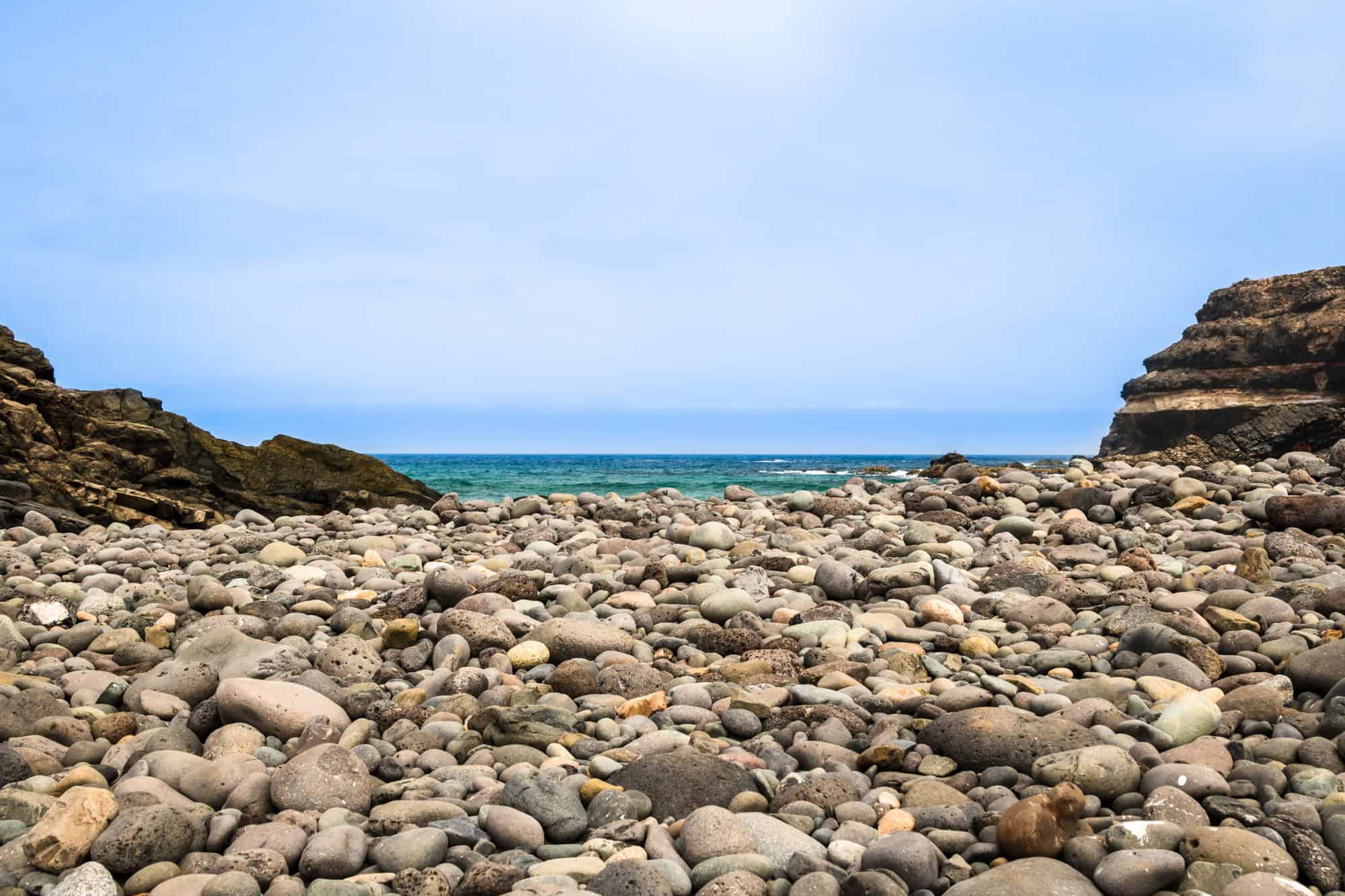 Blick auf einen kiesbedeckten Strand mit Felsen und Meer im Hintergrund, typisch für spanische Küsten.